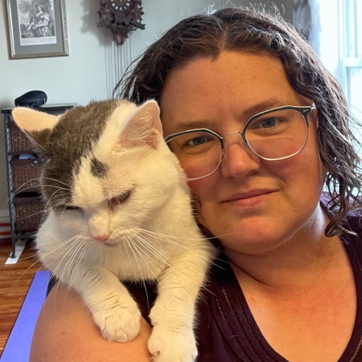 A white woman looks straight at the camera with a half-smile. She has curly brown hair and glasses. There's a soft white-and-grey cat draped over her shoulder.