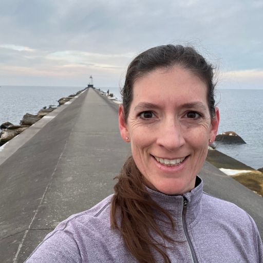 Casual selfie of a white woman with long brunette hair and brown eyes taken in the middle of a run on a pier with Lake Michigan in the background