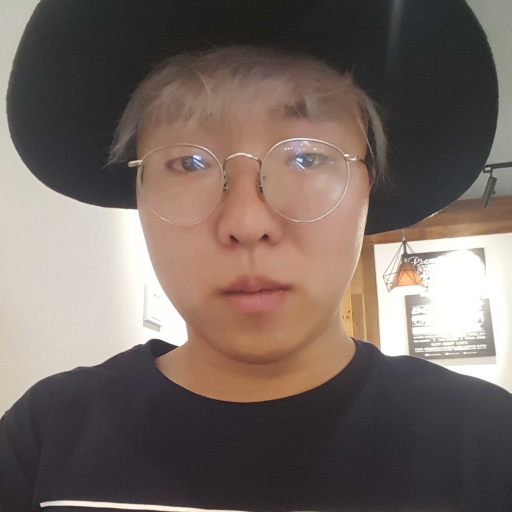 A young Han Chinese person wearing a black shirt and wire-rimmed glasses gazes forwards with a neutral expression. A white fringe of bleached hair peaks out from underneath a black wide-brimmed hat.