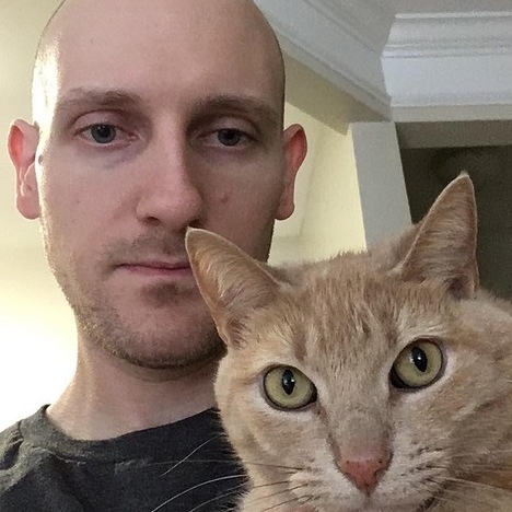Close-up of Jacob, a white, bald man with a light beard, holding his equally-stoic orange tabby cat named Tree.
